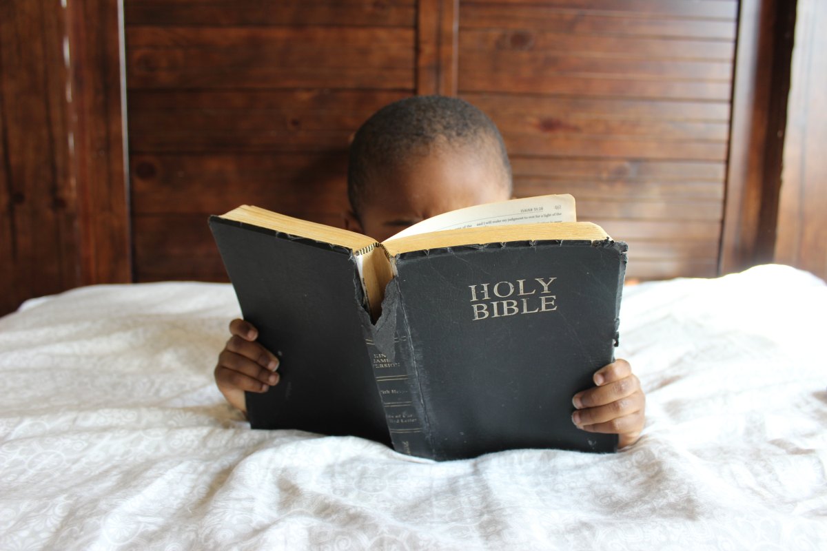 Child reading tattered Bible in bed.